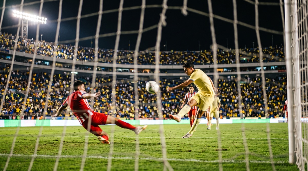 Delantero de amarillo remata ante un defensa de rojo y blanco en un estadio lleno