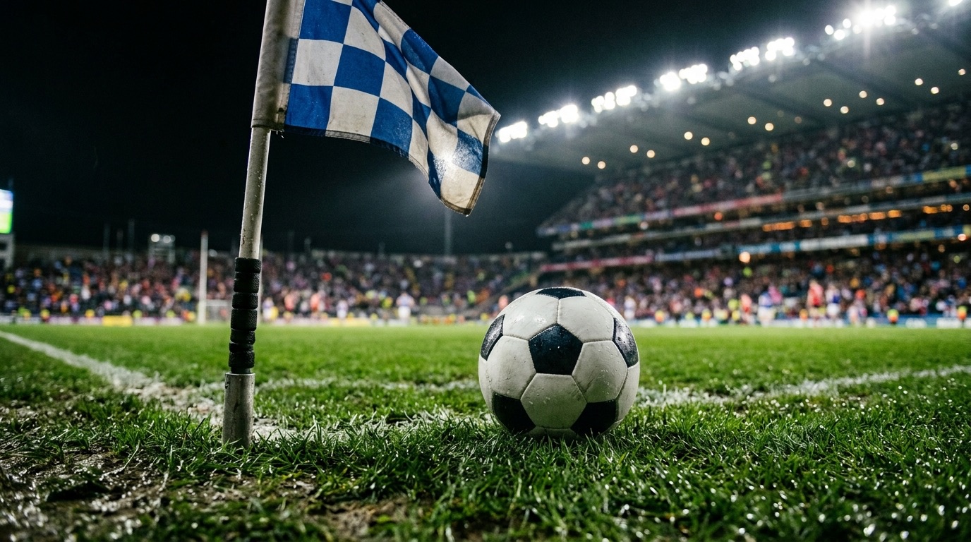 Balón junto al banderín de córner a cuadros azules y blancos, de noche, con la tribuna iluminada al fondo